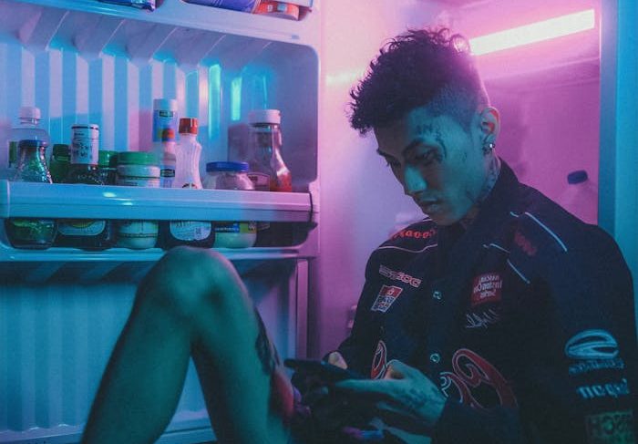 Tattooed man uses phone while sitting beside neon-lit open fridge.